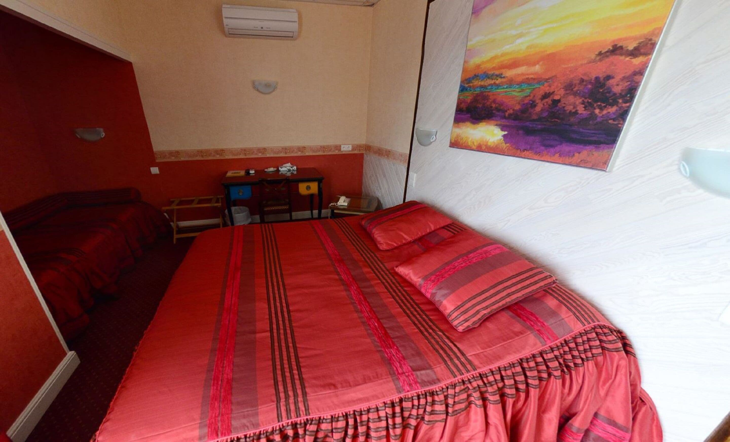 Une chambre d’hôtel avec un grand lit, un bureau et une chaise. Il y a un tableau au mur et un appareil de climatisation au plafond.