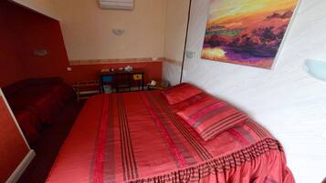 Une chambre d’hôtel avec un grand lit, un bureau et une chaise. Il y a un tableau au mur et un appareil de climatisation au plafond.