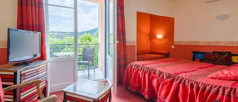 A hotel room with a bed, a TV, a desk, and a chair. There is a balcony with a view of greenery.