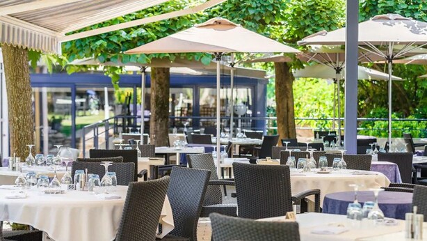 Un área de comedor al aire libre con mesas y sillas, cubierta por sombrillas y rodeada de vegetación.