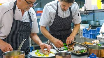Two chefs preparing food in a kitchen with stainless steel pots and a blue plastic container.