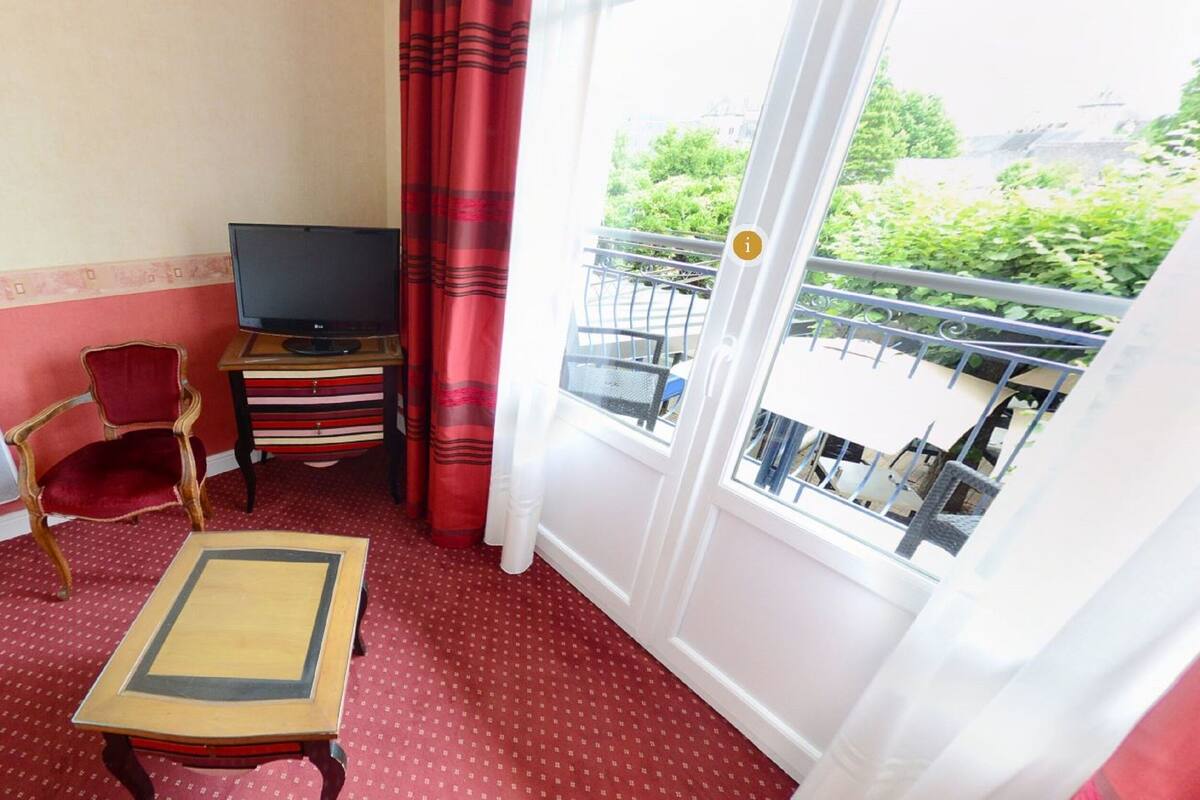 A small room with a red carpet, a TV on a wooden stand, a red chair, a small table, and a balcony with a view of greenery.