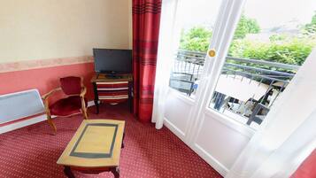 A small room with a red carpet, a TV on a wooden stand, a red chair, a small table, and a balcony with a view of greenery.