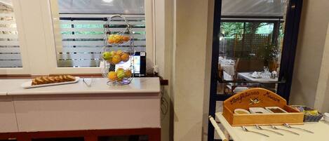 Ein Küchenbereich mit Obstkorb auf der Arbeitsfläche, einem Tisch mit Besteck und einem Fenster mit Jalousien.