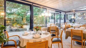 Un restaurante con mesas redondas y sillas de madera, ventanas amplias con vistas a zonas verdes y piso de baldosas.