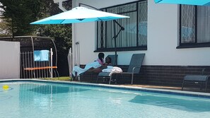 Un'area piscina con una poltrona da sdraio e un ombrellone, un'area giochi recintata e un edificio con finestre e un muro in mattoni.