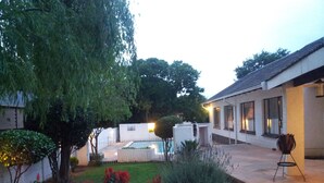 Un entorno residencial con piscina, una casa blanca y un jardín con diversas plantas.