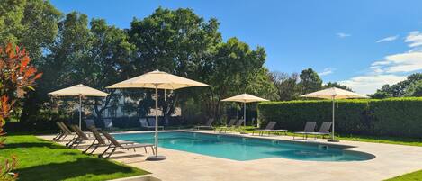 Piscina rectangular con sillones beige y sombrillas, rodeada por un césped cuidado y setos.
