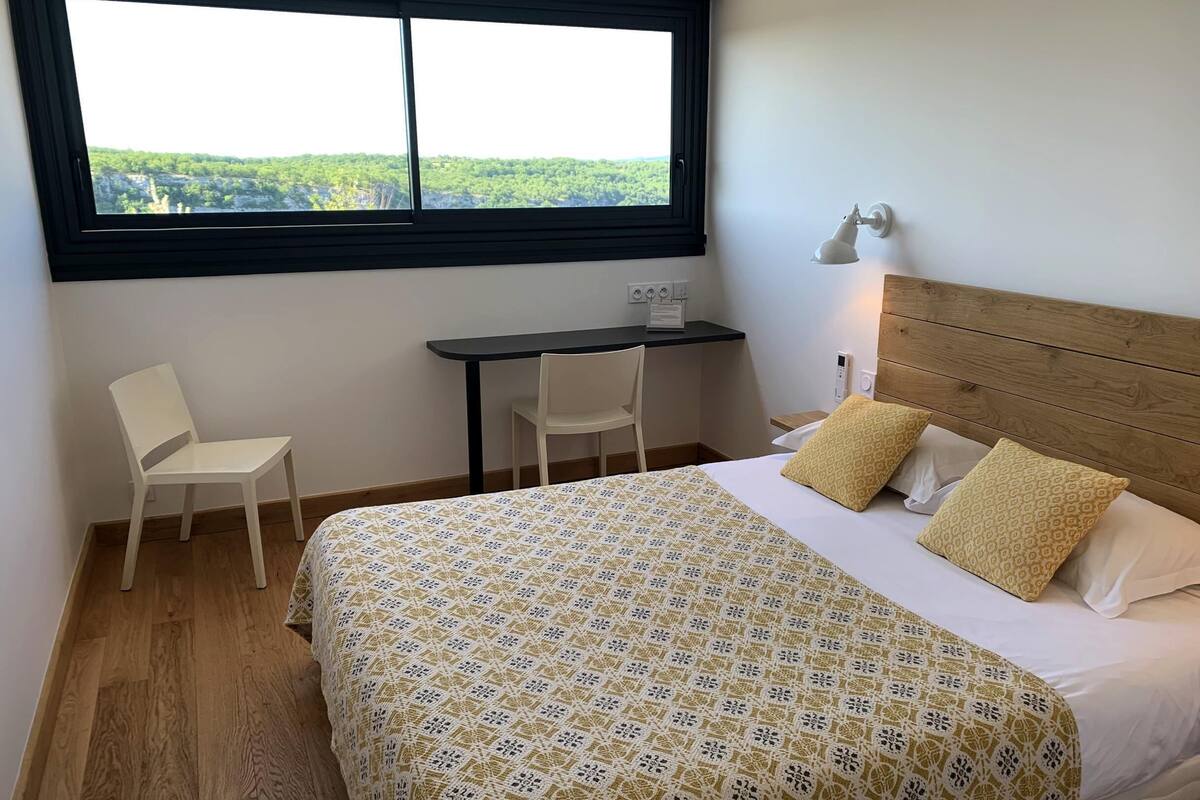 A bedroom with a bed, a desk, two chairs, a window with a view, and a bedside lamp.