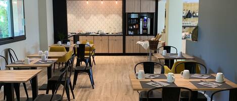 A modern dining area with wooden tables and black chairs, a kitchen in the background, and a patterned wall.