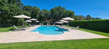 Una piscina con sillas de descanso y sombrillas, rodeada de césped y árboles bien cuidados.