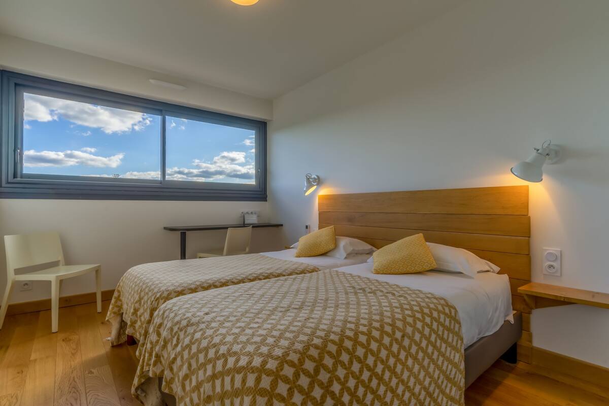 A bedroom with two beds, a desk, a chair, and a window with a view of the sky.