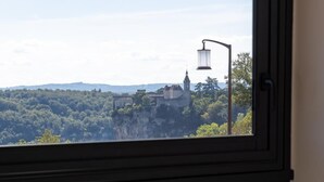A room with a window overlooking a cityscape, a small table with a vase, and a chair. - Hotel Bellaroc (Rocamadour)