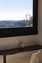 Una habitación con ventana que da a una vista urbana, una mesita con un florero y una silla.
