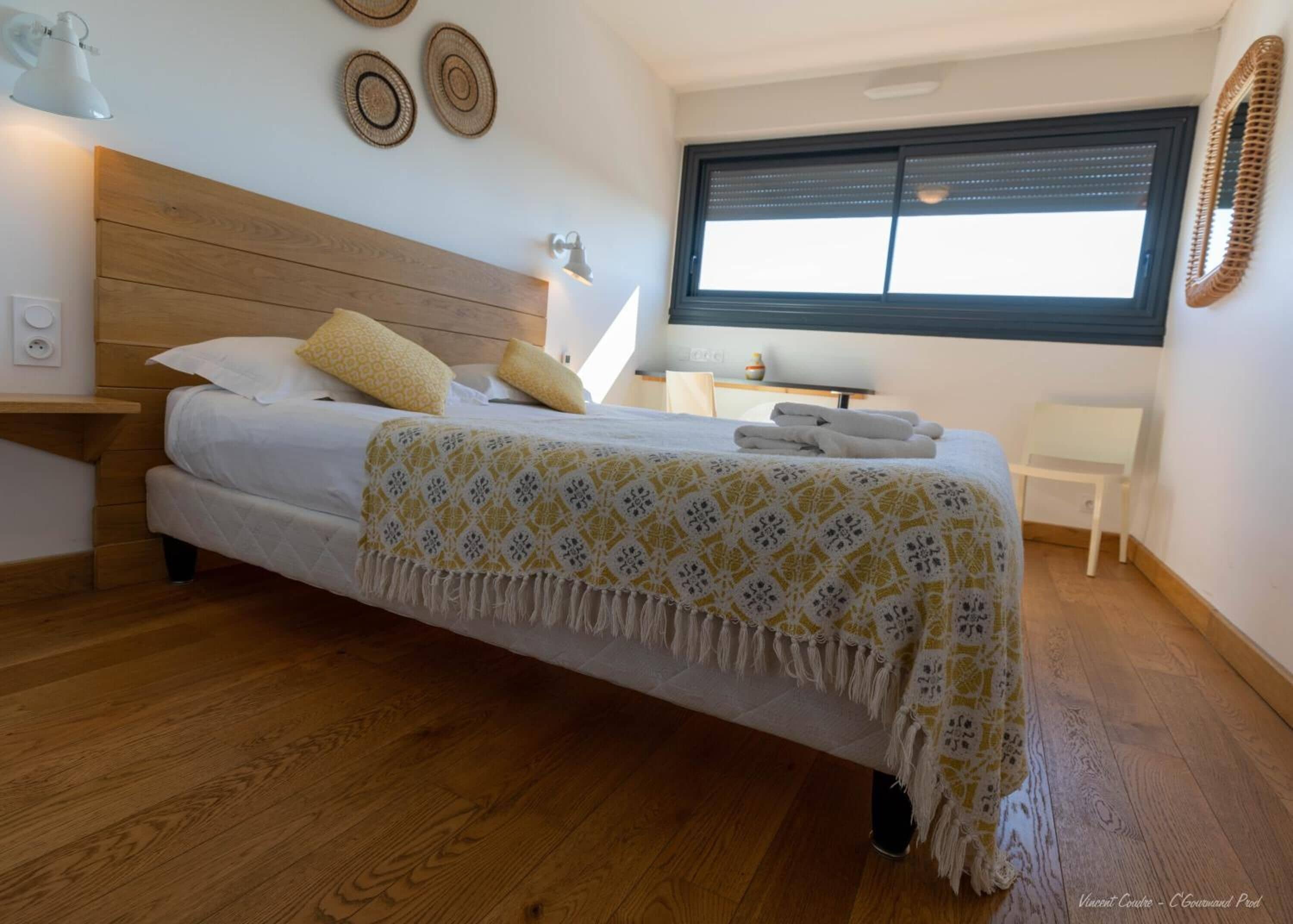 A bedroom with a wooden headboard, a bed with white bedding and yellow pillows, a bedside table with a lamp, a small desk with a chair, and a window with blinds.
