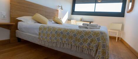 A bedroom with a wooden headboard, a bed with white bedding and yellow pillows, a bedside table with a lamp, a small desk with a chair, and a window with blinds.