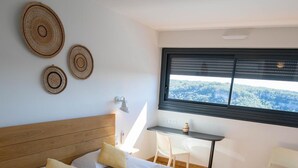 A bedroom with a wooden headboard, a bed with a patterned bedspread, two pillows, a bedside table with a lamp, and a window with a view of a forest. - Hotel Bellaroc (Rocamadour)