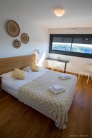 Un dormitorio con cabecera de madera, cama con colcha estampada, dos almohadas, una mesita de noche con lámpara, y una ventana con vistas a un bosque.