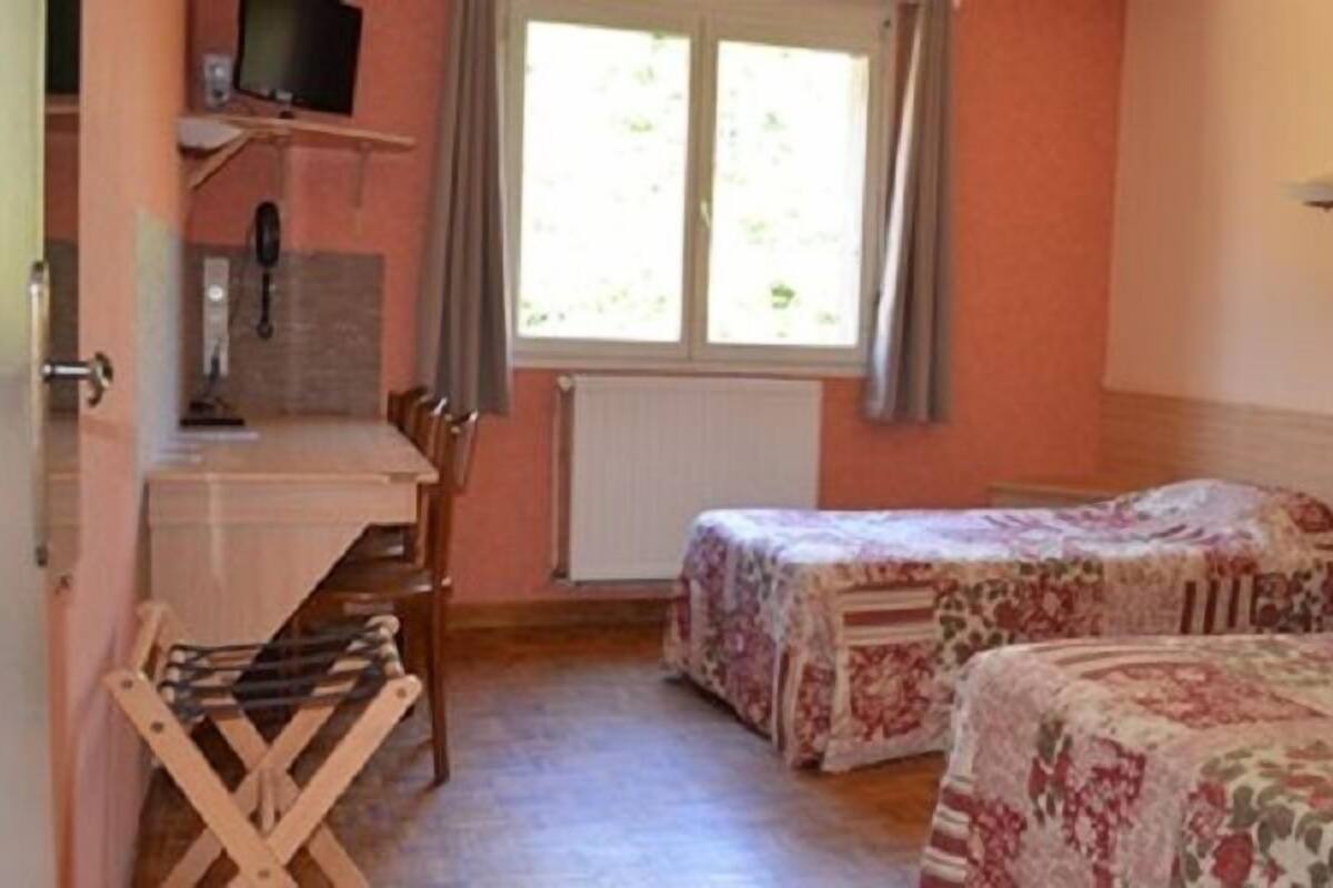 Ein Zimmer mit zwei Betten, einem Schreibtisch und einem Sessel. Es gibt ein Fenster mit Vorhängen, eine Heizung und einen Fernseher auf einem Regal.