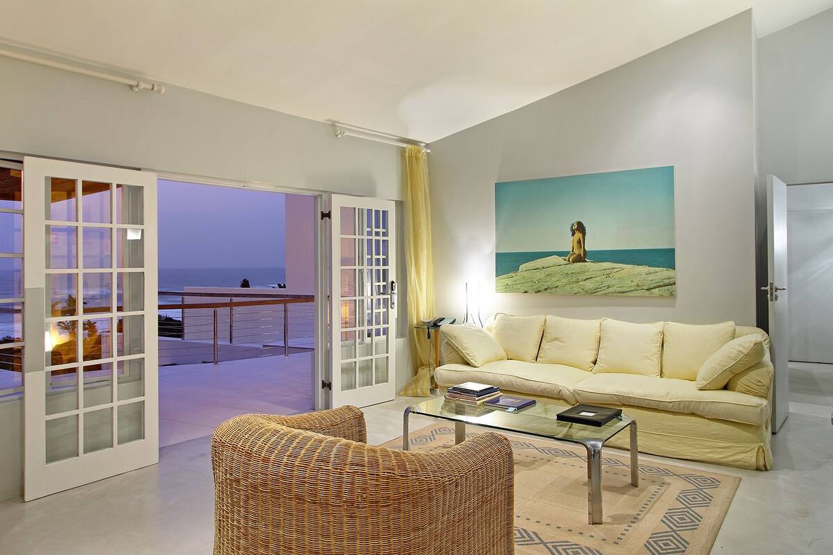 A living room with a sofa, two wicker chairs, a glass coffee table, and a painting of a beach scene.
