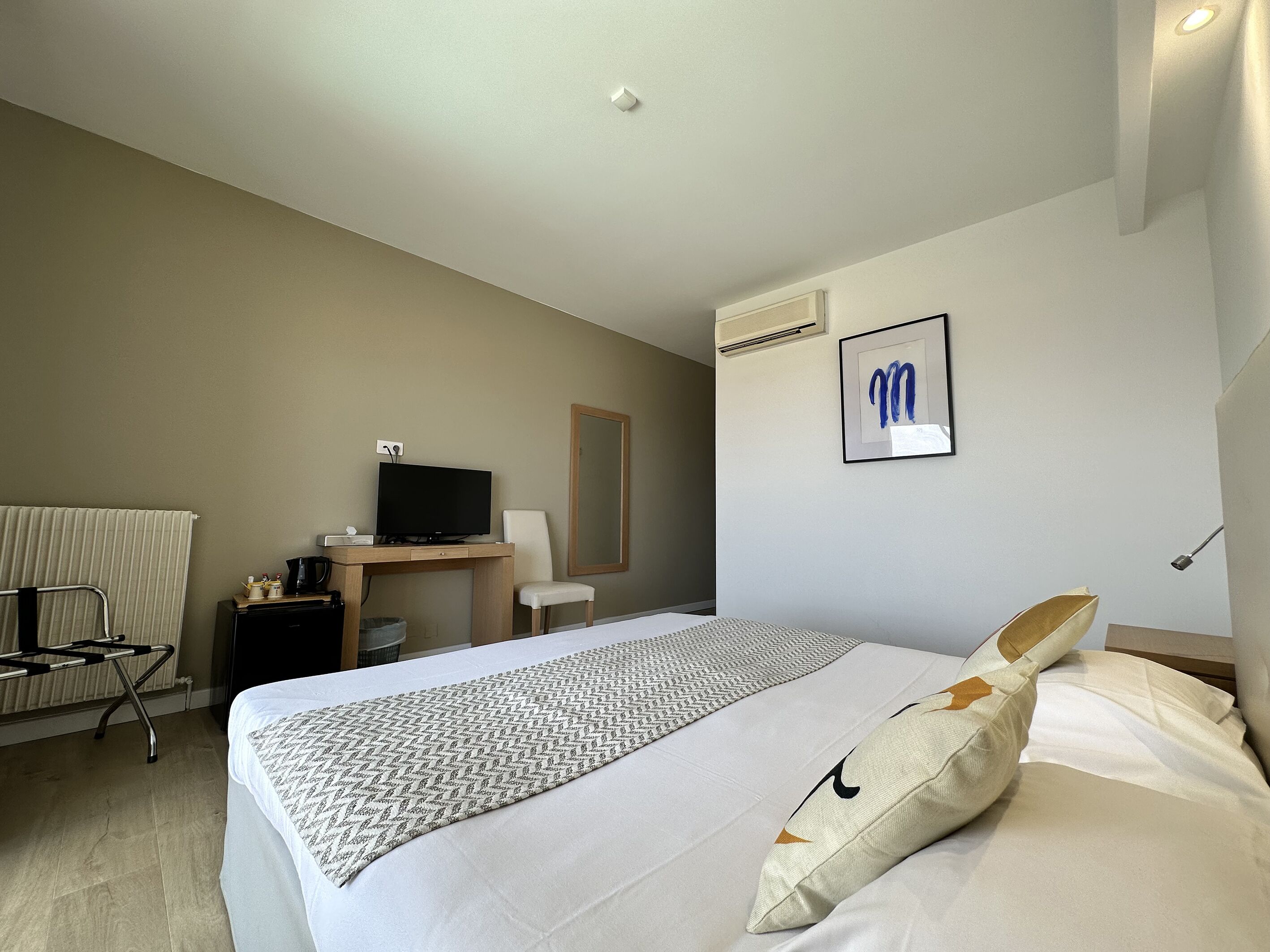 A hotel room with a bed, a desk with a television, a chair, a radiator, and a framed artwork on the wall.