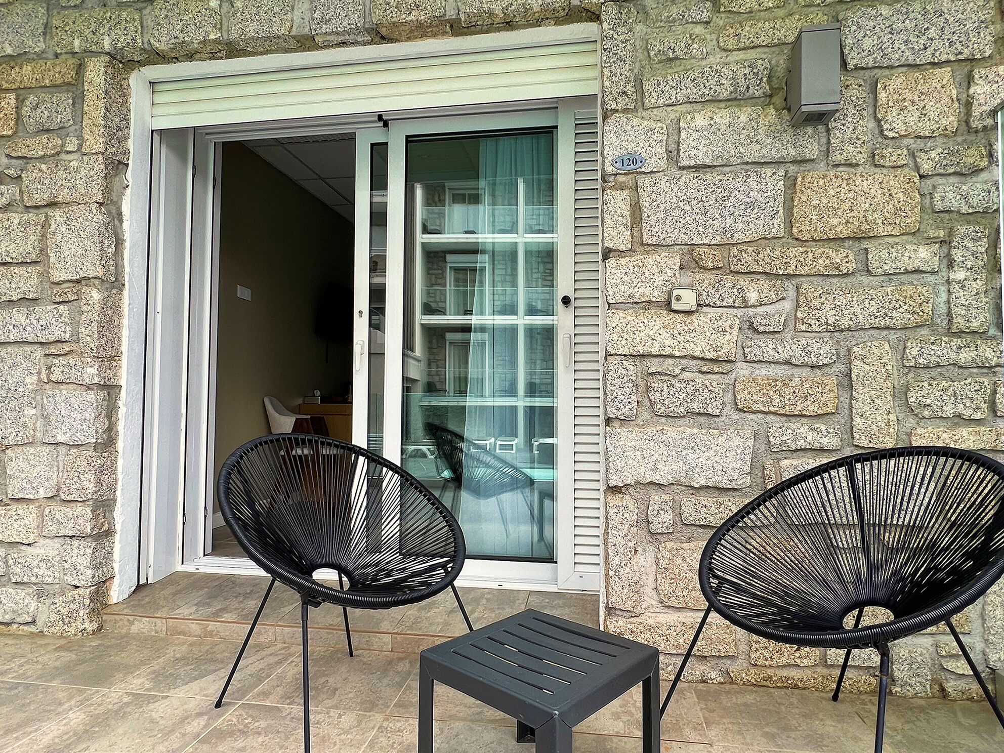 A stone building with a sliding glass door leading to a balcony with two black chairs and a small table.
