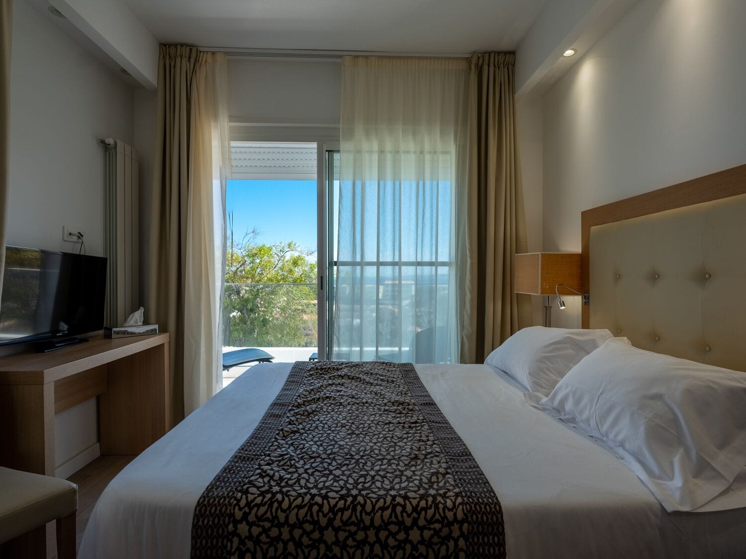 A hotel room with a bed, a TV, a desk, and a balcony with a view.