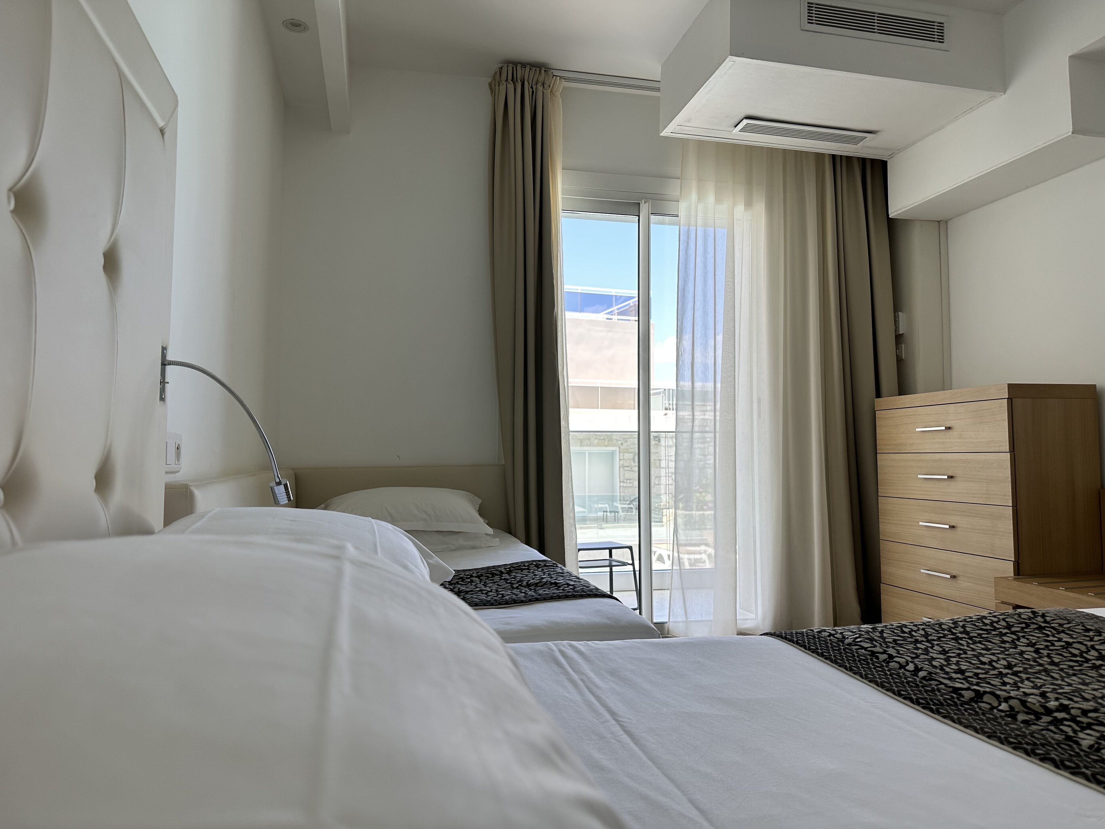 Habitació d'hotel amb dos llits, un taulell de fusta, bany amb dutxa i un balcó amb cortines.