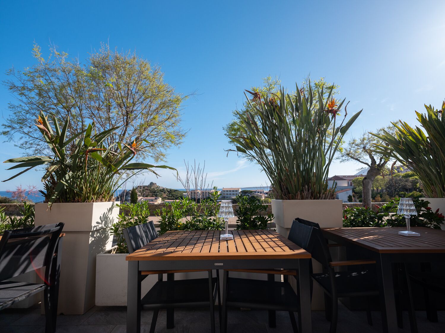 An outdoor patio with a wooden table and chairs, surrounded by potted plants and overlooking a scenic view.