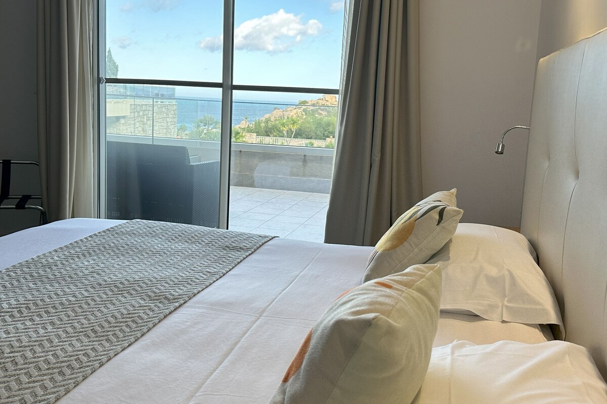 A hotel room with a large window, a bed with white linens, and a view of the city and hills.