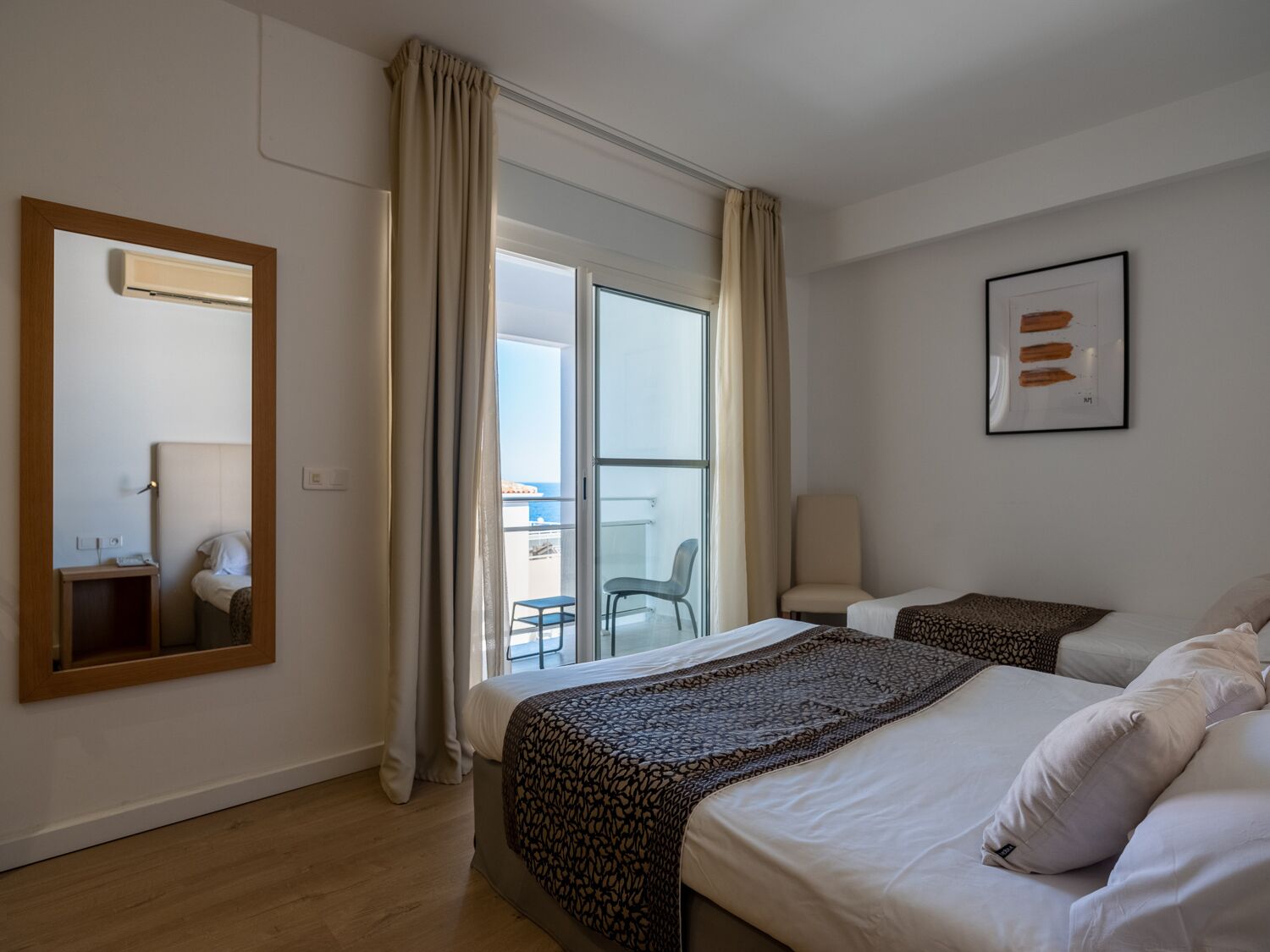 A hotel room with a large bed, a smaller bed, a balcony with a chair, a mirror, and a framed picture on the wall.