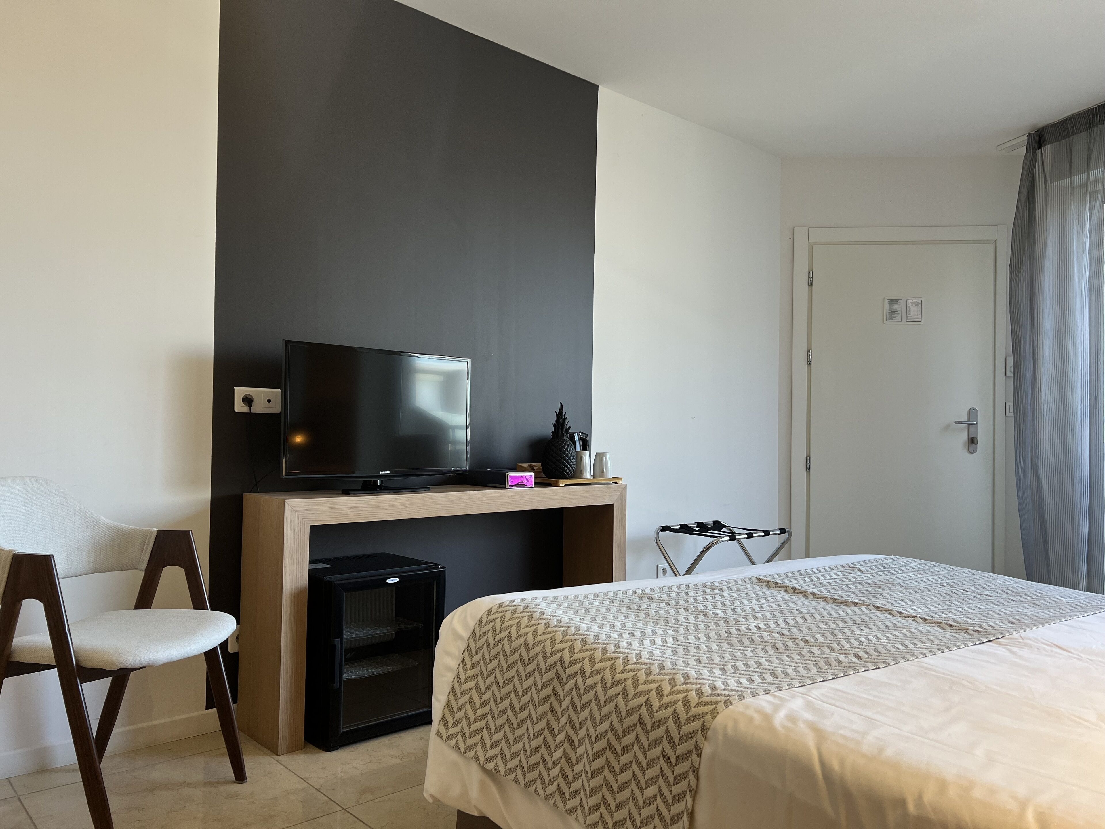 A hotel room with a bed, a TV on a wooden stand, a small fridge, a chair, and a door with a curtain.