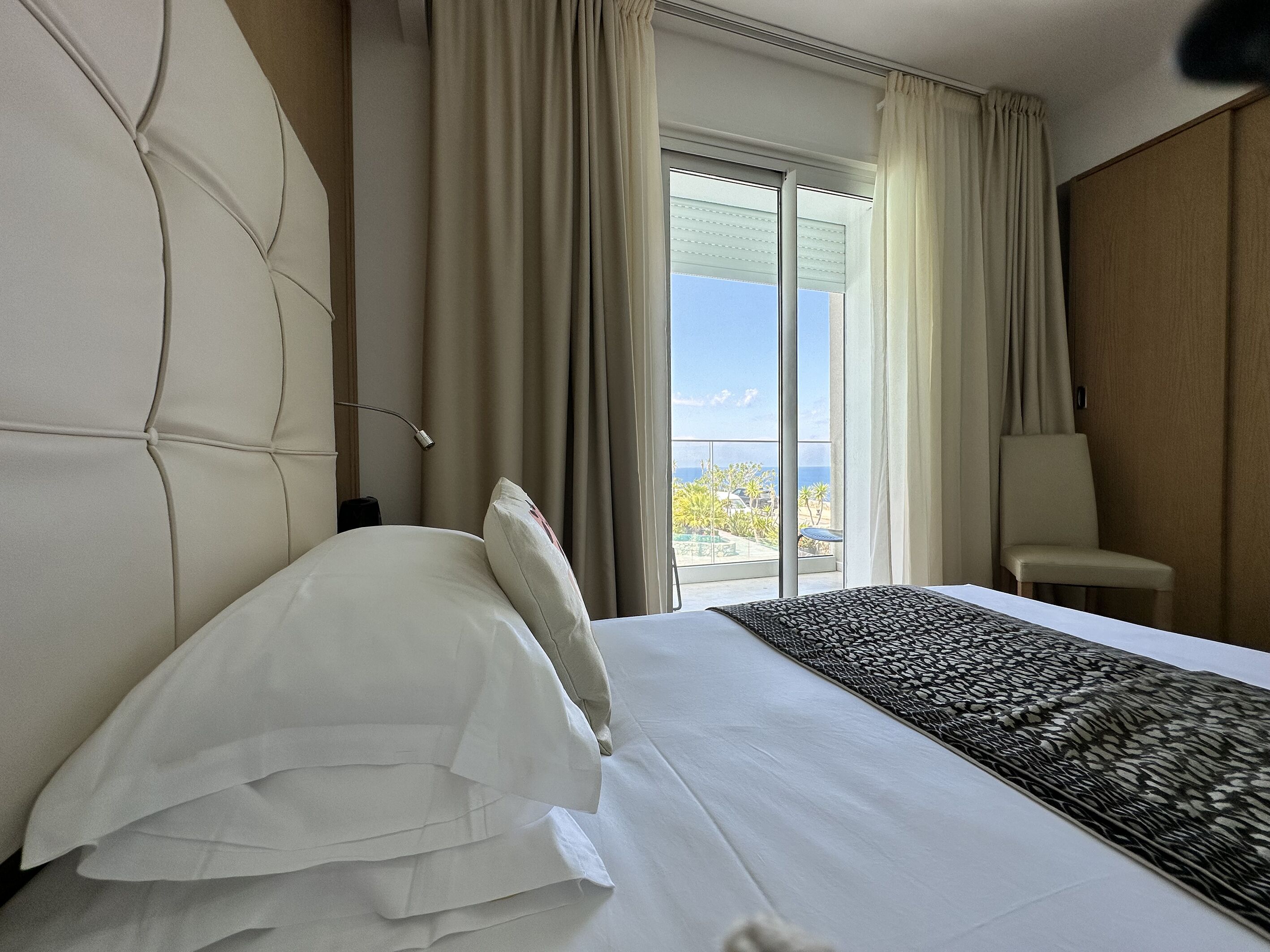 A hotel room with a large bed, a chair, a window with a view of the outdoors, and a curtain.