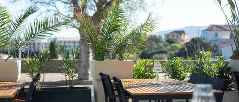 An outdoor seating area with wooden tables and chairs, surrounded by potted plants and trees.