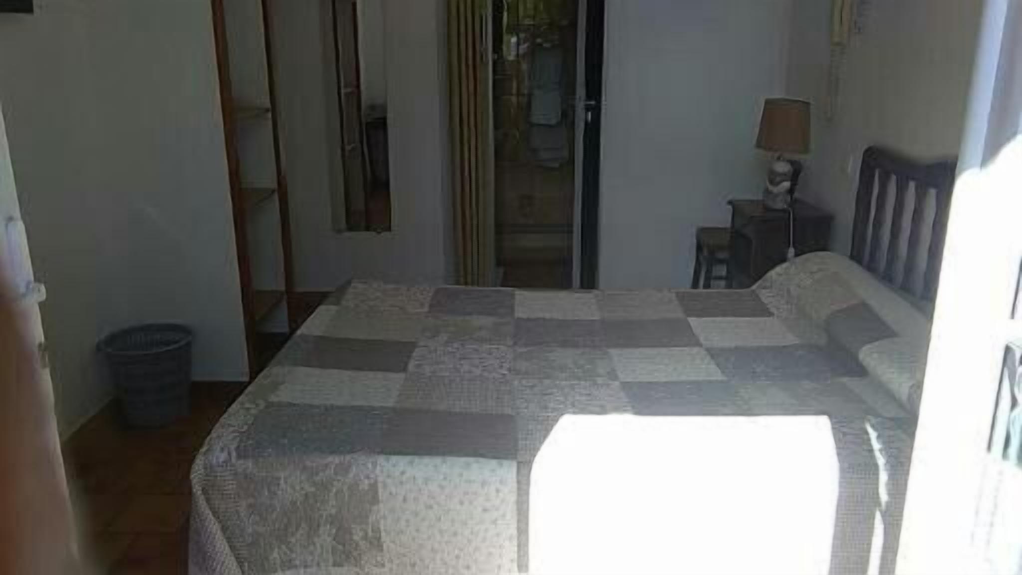 A bedroom with a bed, a small table, a chair, a lamp, and a trash bin.