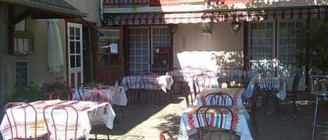 Un área de comedor al aire libre con mesas y sillas, un edificio con balcón y un jardín.