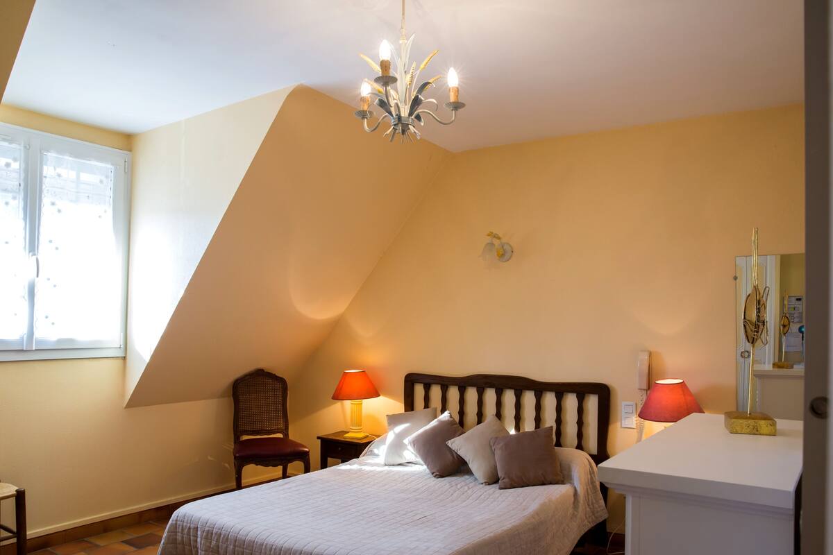 A bedroom with a sloped ceiling, a bed with a wooden headboard, two bedside lamps, a chair, and a small table.