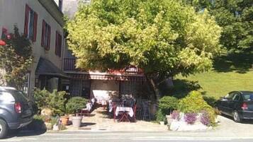A building with a covered outdoor seating area, a car parked on the street, and a tree in front.