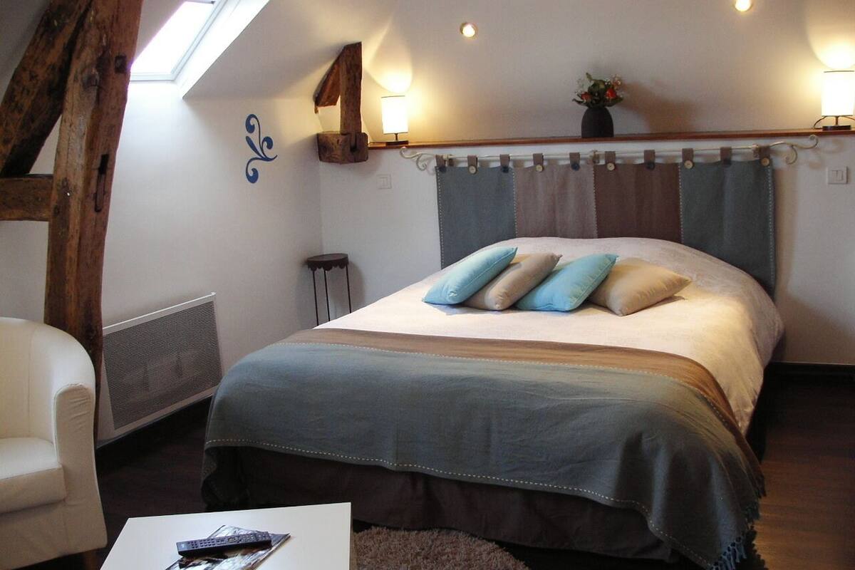 A bedroom with a bed, a chair, a table with a remote control, a window, and a wall-mounted lamp.