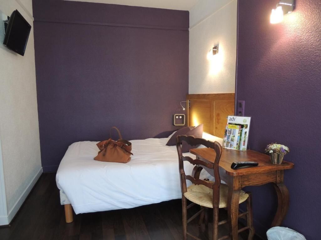 Una habitación de hotel con una cama, una silla de madera, una mesita pequeña, un control remoto, una revista y un florero con flores.