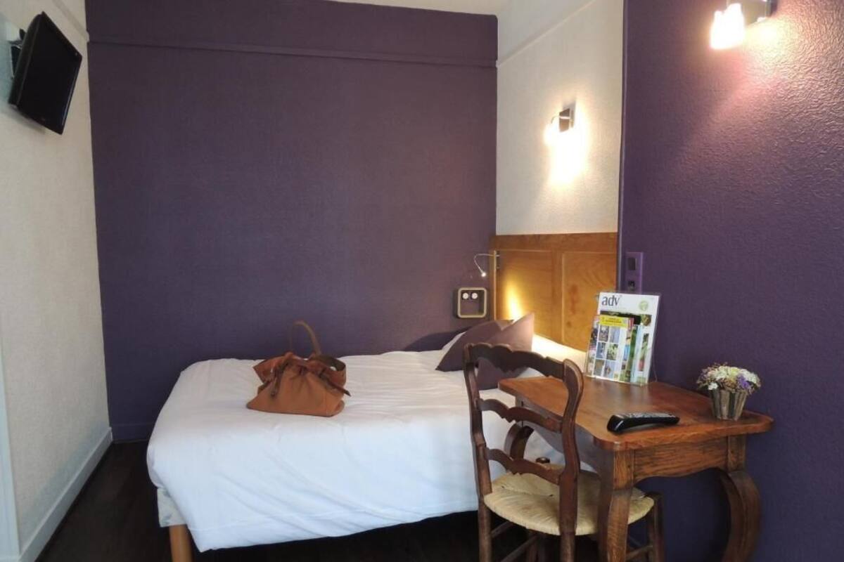A hotel room with a bed, a wooden chair, a small table, a remote control, a magazine, and a vase with flowers.