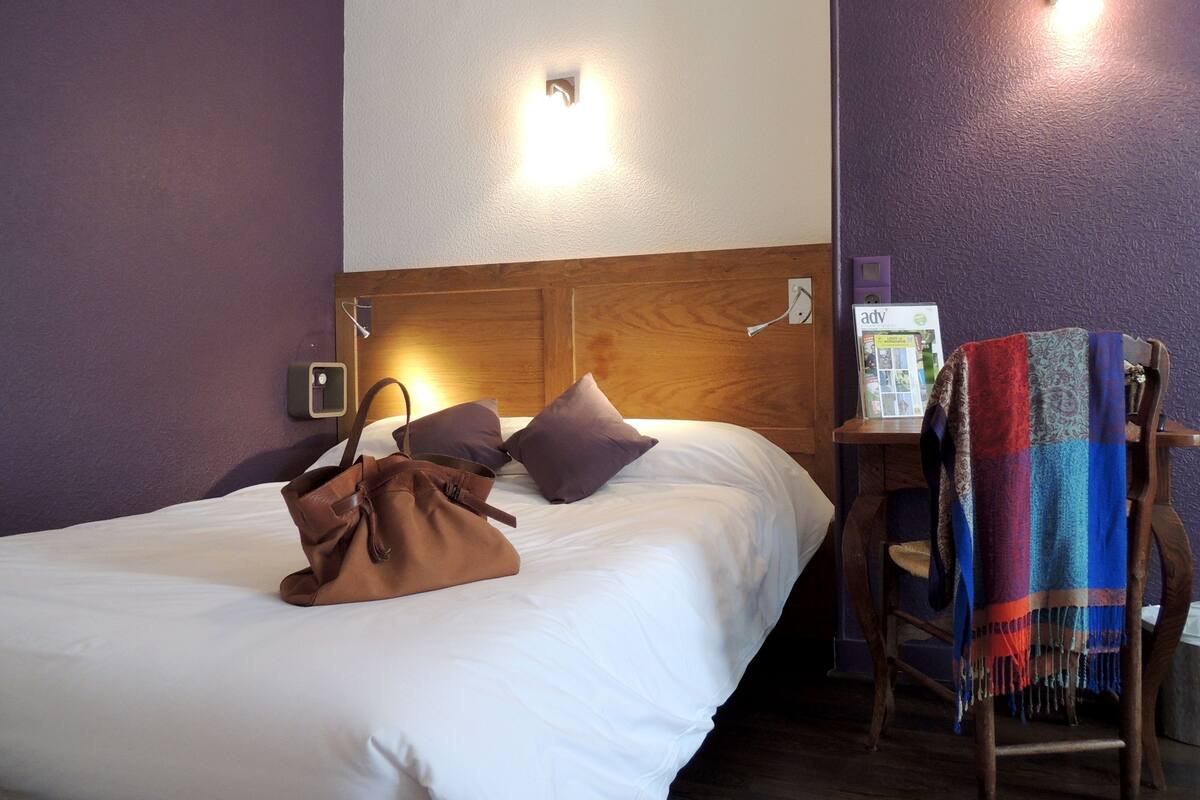 A hotel room with a wooden headboard, a bed with white bedding, a brown handbag on the bed, a chair with a colorful blanket, and a magazine on a small table.