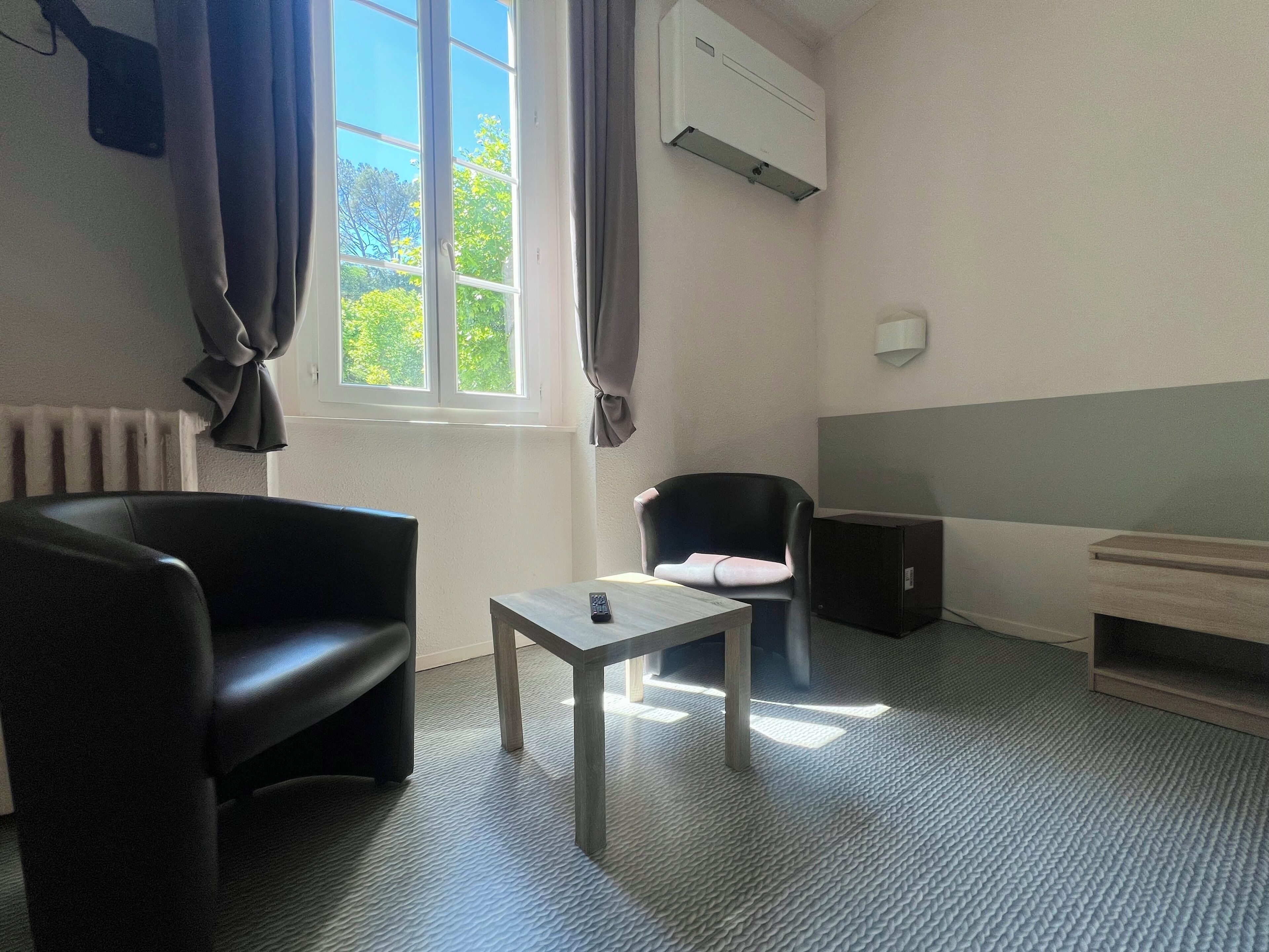 A room with a black chair, a small table with a remote, an air conditioner, a radiator, and a window with curtains.