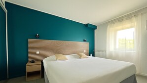 A hotel room with a large bed, a nightstand, a wall-mounted lamp, and a window with curtains. -  Logis Hôtel des Thermes  (Lamalou-les-Bains)