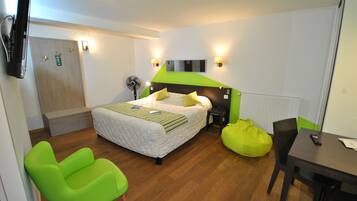 Una camera d'albergo con un letto, una scrivania, una sedia, un pouf verde, una ventola da pavimento e un televisore fissato alla parete.