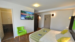 A hotel room with a bed, a green chair, a television on the wall, and a wooden door. - Hostellerie du Perigord Vert (Brantôme en Périgord)