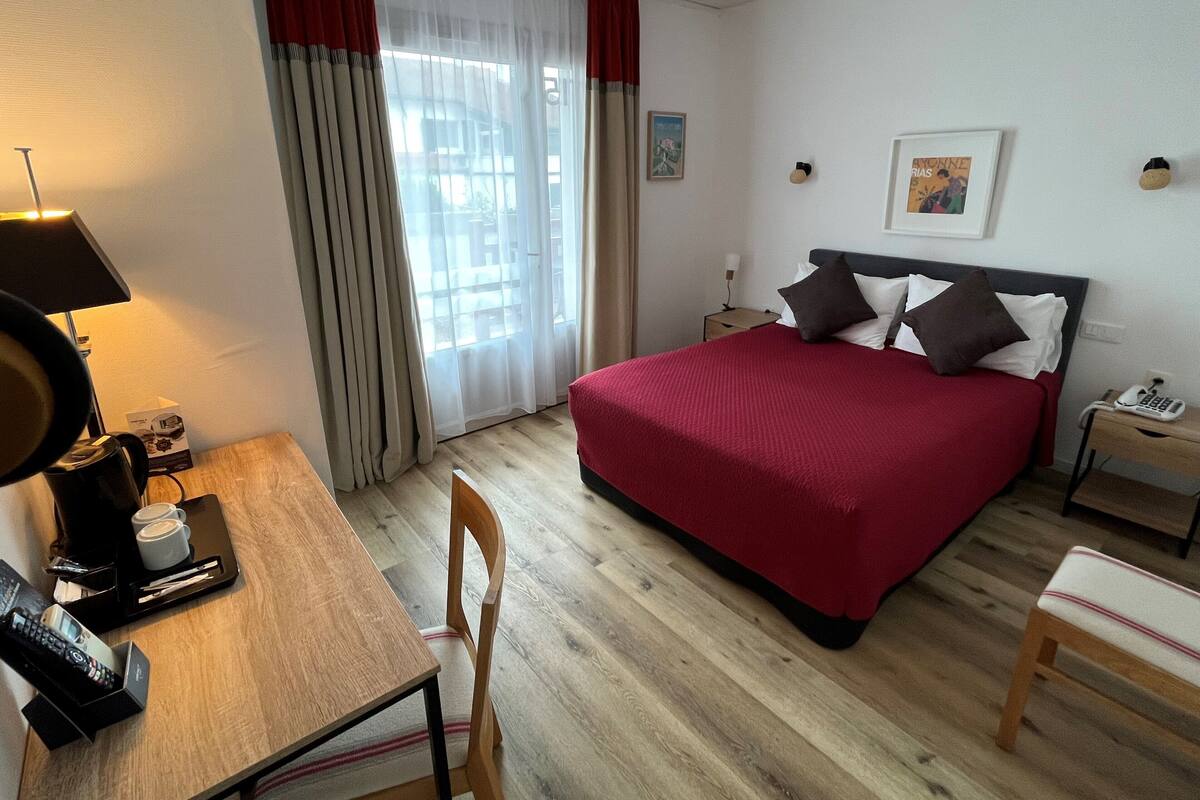 Een hotelkamer met een bed, een bureau met een lamp, een stoel, een nachtkastje met een telefoon en een raam met gordijnen.