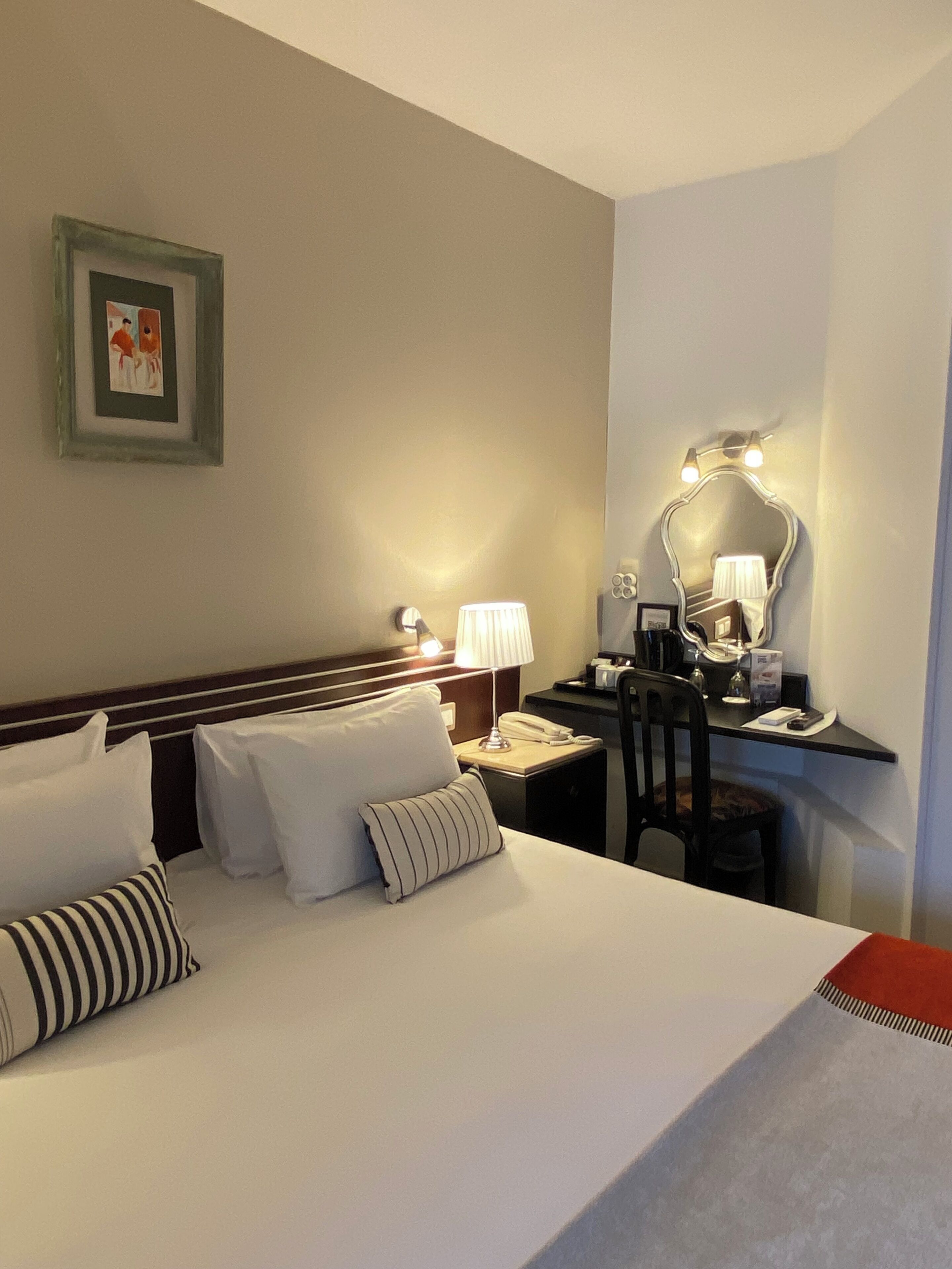 A hotel room with a bed, bedside table, lamp, chair, desk, mirror, and a framed picture on the wall.