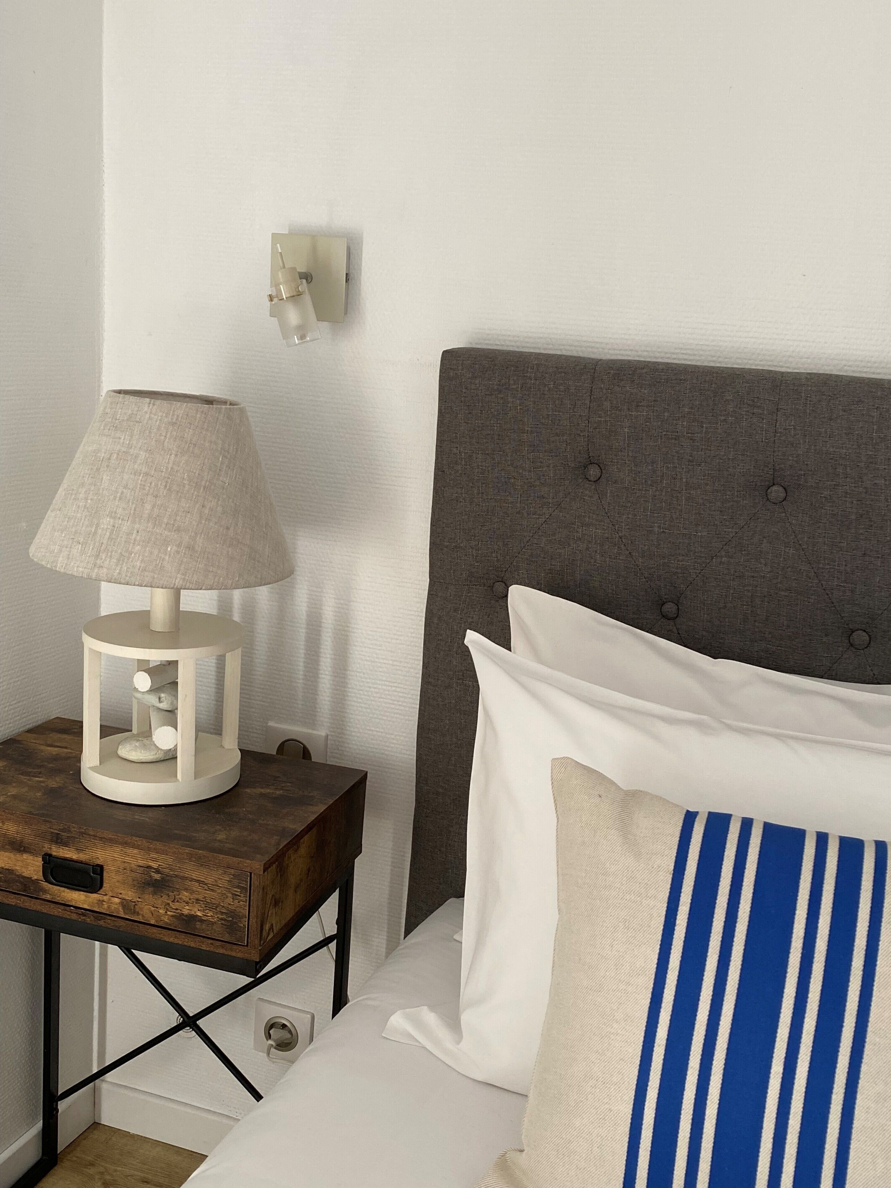 Una cama bien tendida con un cabecero tapizado en gris, una mesita de noche con una lámpara y un aplique de pared.