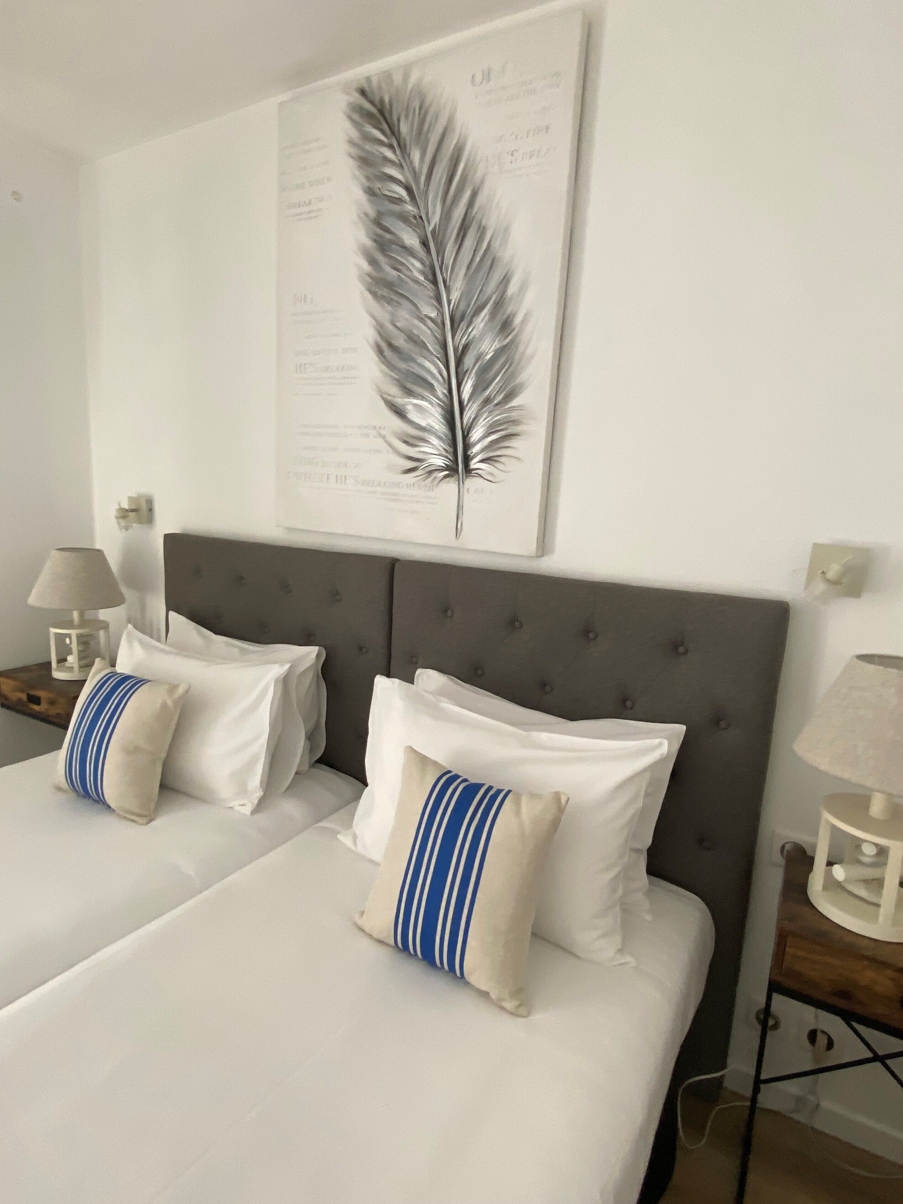 A hotel room with two beds, a grey upholstered headboard, white bedding, and a feather artwork on the wall.